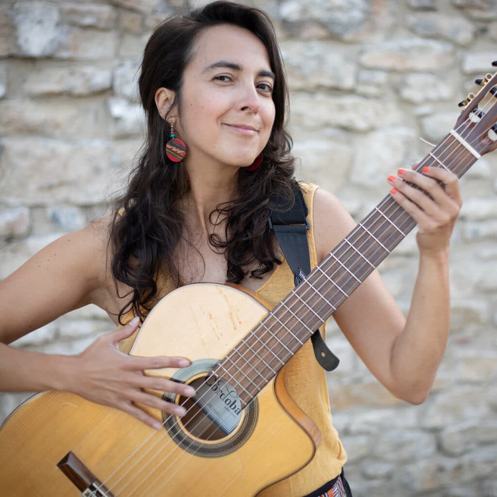 Woman with long dark hair holding guitar in front of stone wall