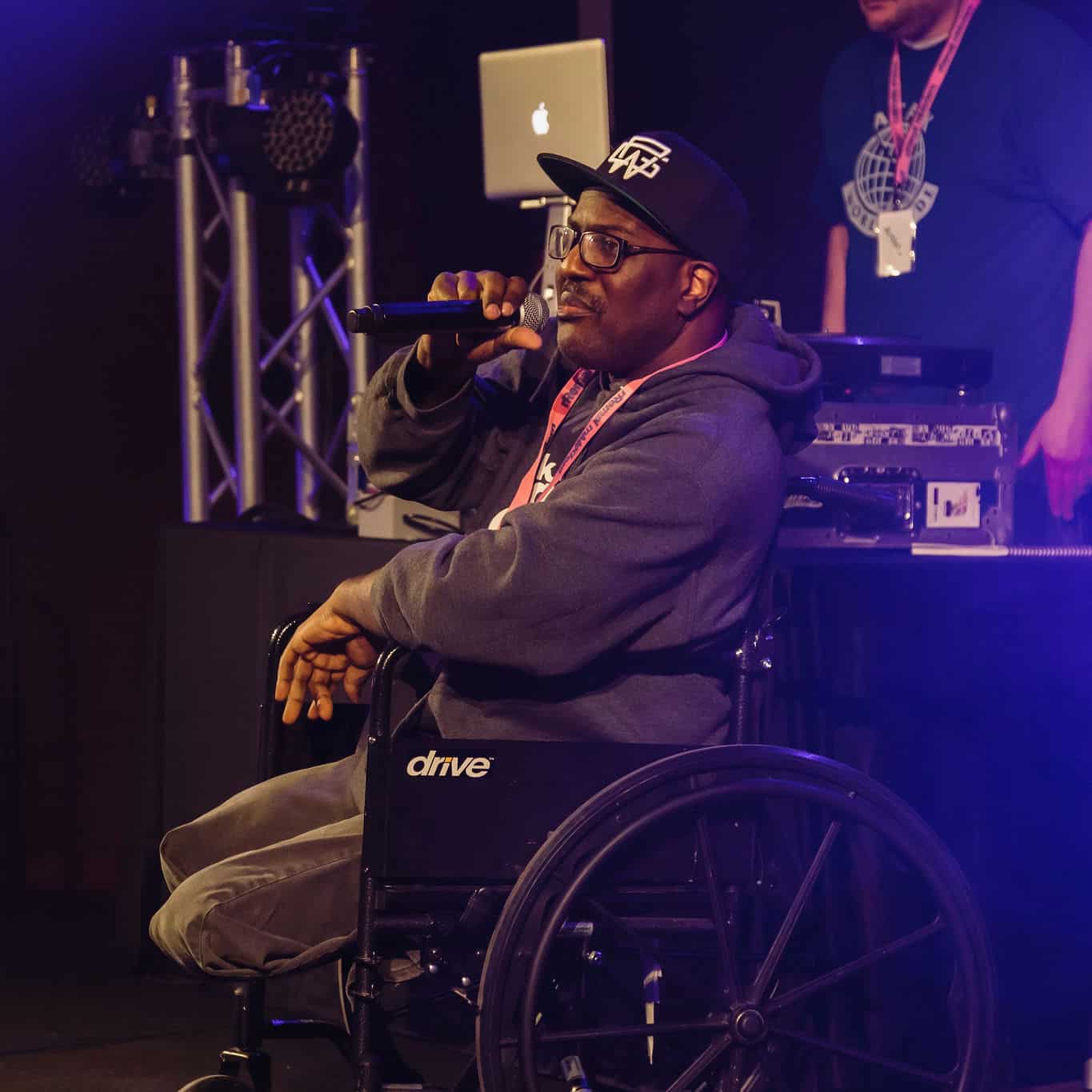 Man in wheelchair wearing baseball cap holding microphone to face. DJ in background.
