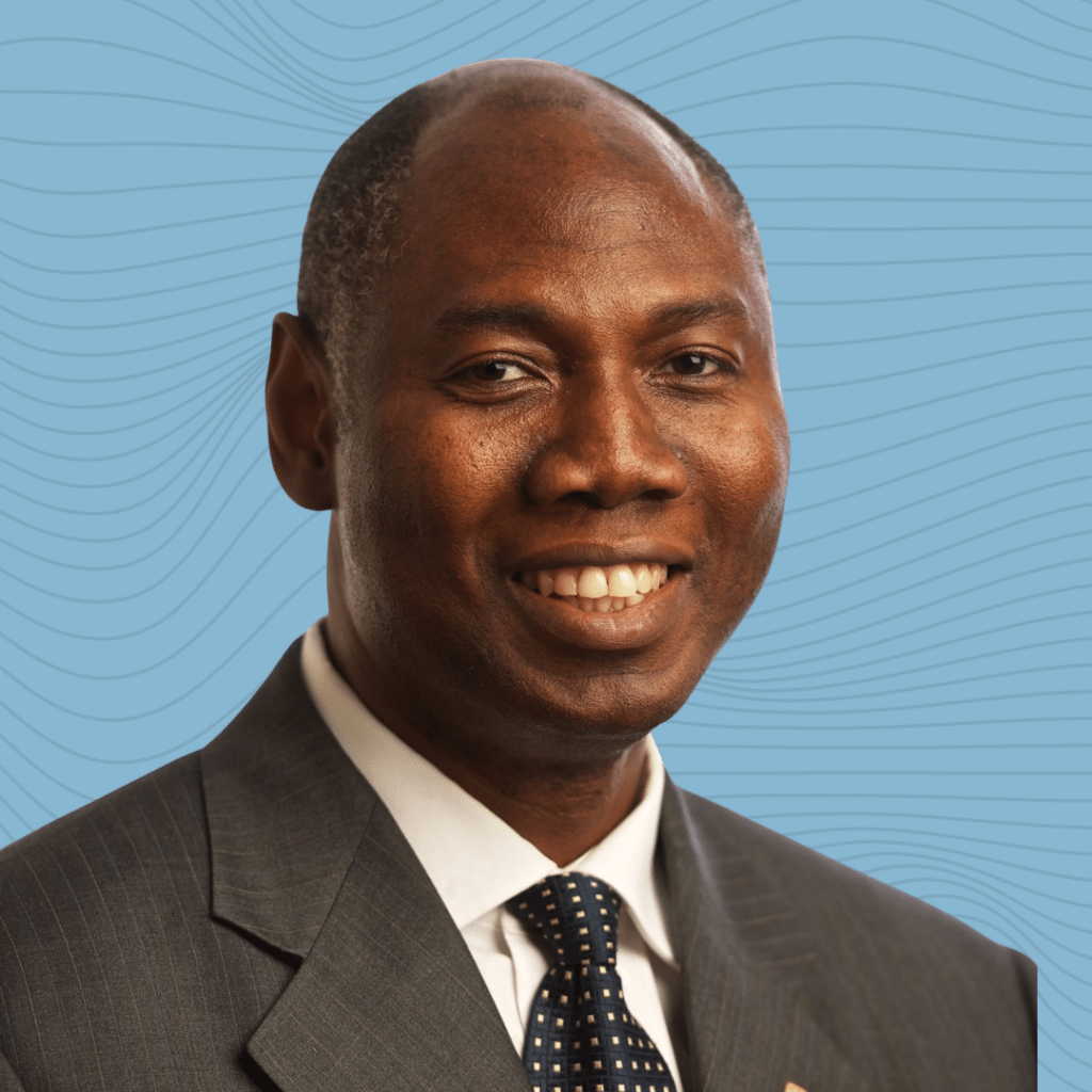 A black man is smiling towards the camera, dressed in a grey suit with white shirt and tie.