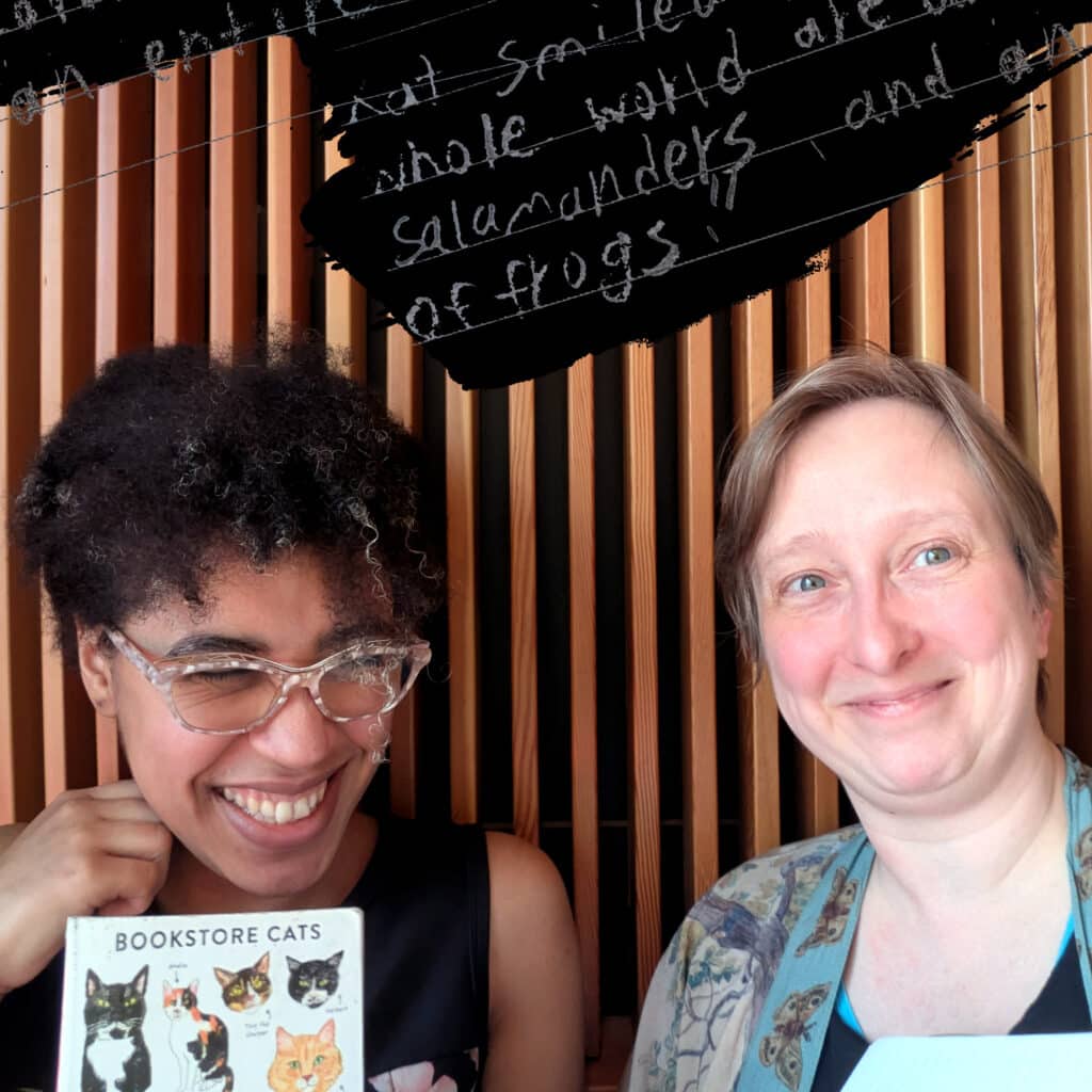 Photo of two people against wood slat wall. Person on left is holding book titled "Bookstore Cats"